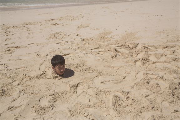 砂浜に埋まり自身の影で方角を確認する男性 ［モデル：大川竜弥］